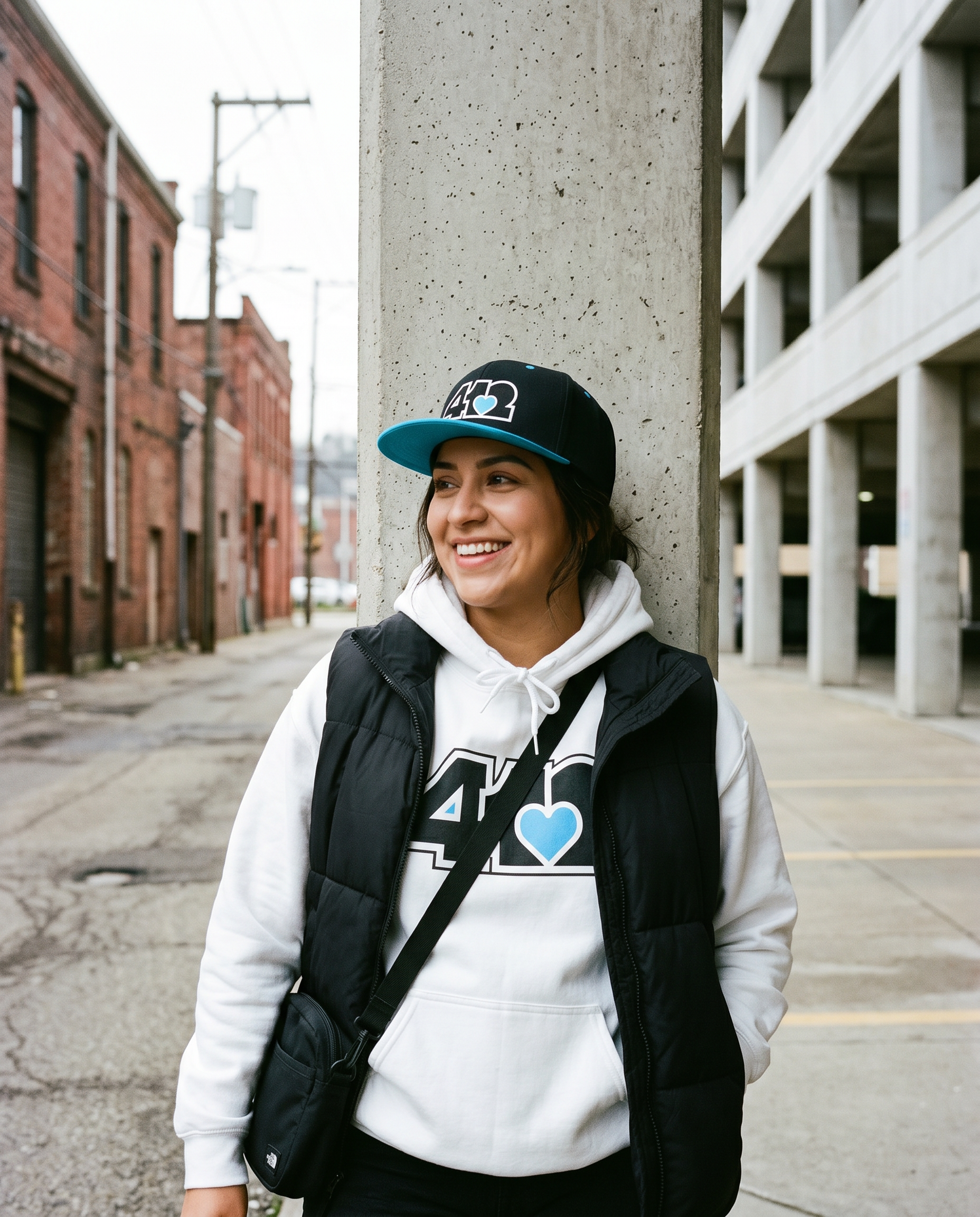 Person wearing a white hoodie with a 4OneLove2 logo, black jacket, and cap, standing against a concrete wall in an urban setting.